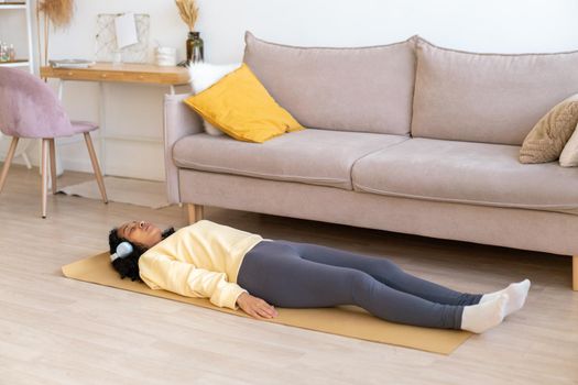 African-american woman wearing sportswear and earphones lying on mat in living room. Having rest after doing stretching. Listening to music while exercising at home. Training during quarantine