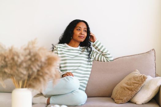 Happy young african-american female sitting on couch in living room. Concept of enjoying life and slow pace lifestyle. Thoughtful smiling woman self-reflecting