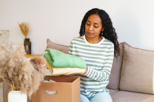 Young african-american female in casual packing clothes for charity donation in cardboard box while sitting on sofa in living room. Concept of shipping stuff due to move. Giving things away