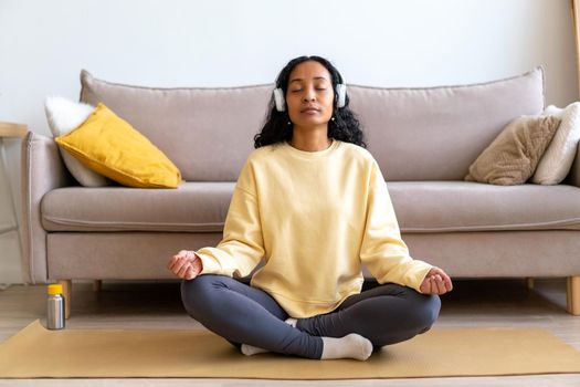 African-american woman in earphone listening to nature sounds while meditating on mat in living room. Concept of mindful living lifestyle. Wellness and wellbeing on breathing therapy at home