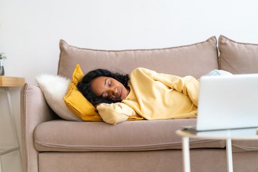 Sleepy young african-american lying and sleeping on sofa in living room while movie playing on laptop. Boring spending free time watching film. Concept of snackable wellness and micro-moment