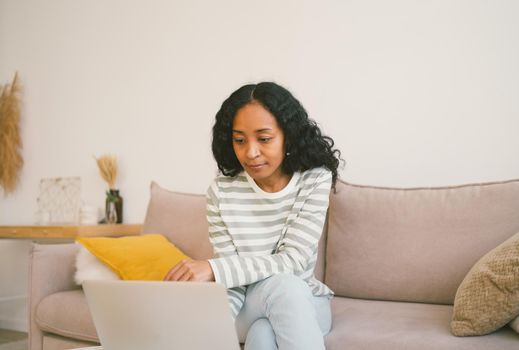 Young african-american female surfing the net on laptop while sitting on couch. Using digital device for remote work and online study. Concentrated and focused woman flexible working from home