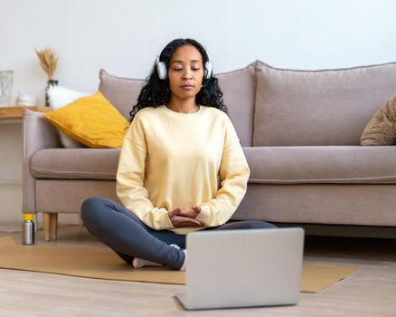 African-american woman sitting on mat in lotus pose while listening to music in earphones and looking at laptop. Doing yoga at home to stress relief. Relaxing and breathing for inner peace