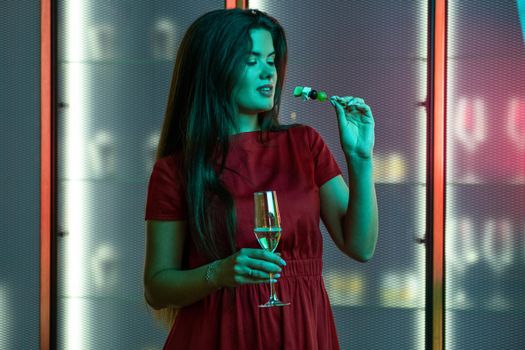 Young long-haired brunette in red dress enjoying champagne with light snacks in nightclub while standing alone in subdued color lighting