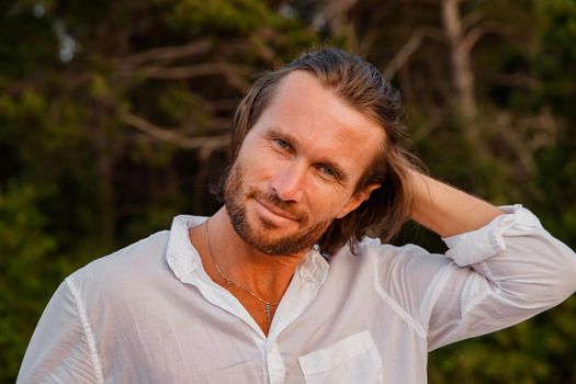 Portrait of handsome brutal man with long hair at sunset, the blue eyes, an easy bristle, he is dressed in a white shirt with a short sleeve, green trees on a background. High quality photo