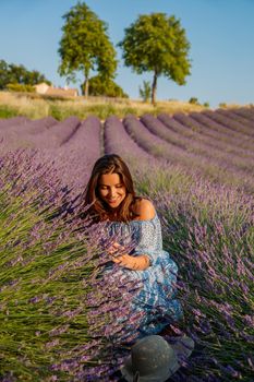 The beautiful young girl in a blue dress sits on the field of a lavender flowers, long curly hair, bright smile, a house of the gardener, trees, perspective of a lavender. High quality photo