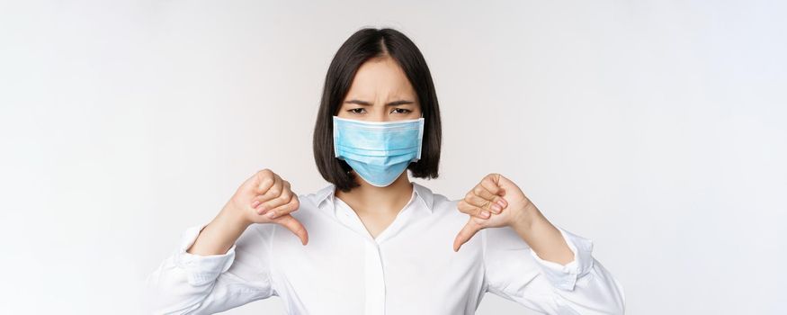 Portrait of asian woman in medical face mask showing thumbs down with disappointed, tired face expression, standing over white background.