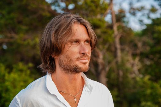 Portrait of handsome brutal man with long hair at sunset, the blue eyes, an easy bristle, he is dressed in a white shirt with a short sleeve, green trees on a background. High quality photo