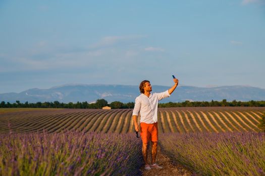 The handsome brutal man with long brunette hair makes selfie in the field of lavender in provence near Valensole at sunset, France, clear sunny weather, in a rows of lavender, red shorts. High quality photo