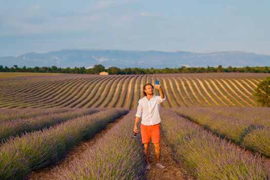 The handsome brutal man with long brunette hair makes selfie in the field of lavender in provence near Valensole at sunset, France, clear sunny weather, in a rows of lavender, red shorts. High quality photo