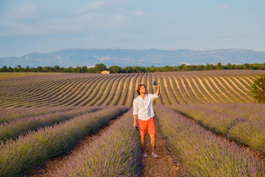 The handsome brutal man with long brunette hair makes selfie in the field of lavender in provence near Valensole at sunset, France, clear sunny weather, in a rows of lavender, red shorts. High quality photo