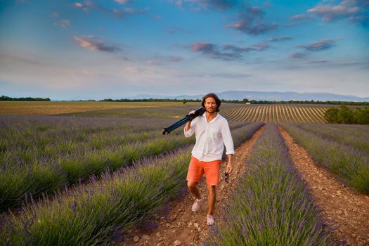The handsome brutal man with long brunette hair is walking with tripod in the field of lavender in provence near Valensole, France, clear sunny weather, in a rows of lavender, red shorts, white shirts. High quality photo