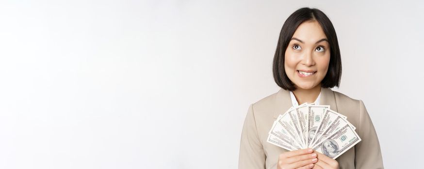 Image of asian corporate woman, happy businesswoman showing money, cash dollars and thinking, standing in suit over white background.