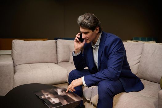 Interior designer in a business suit consults a client on the phone, sitting on the couch and leafing through a catalog with an assortment of upholstered furniture. Home improvement concept