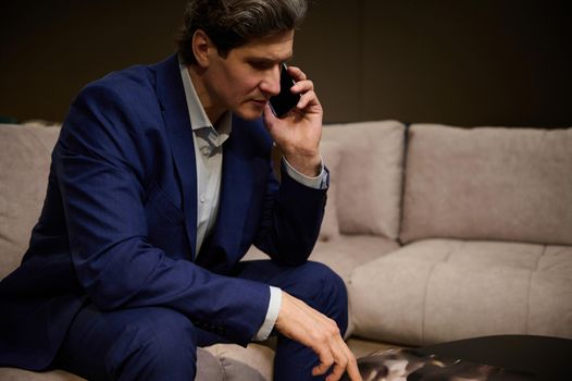 Handsome mature man in business suit looking furniture catalogue and talking on mobile phone, sitting on a comfortable modern velour sofa in a furniture store showroom. Small business, interior design