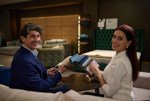 Charming middle aged couple sitting on the sofa in a furniture store, holding a color fabric samples and picking couch seat cover.