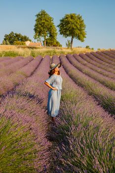 The beautiful young girl in a blue dress and cap walks across the field of a lavender, long curly hair, smile, pleasure, a house of the gardener in the background, trees, perspective of a lavender. High quality photo