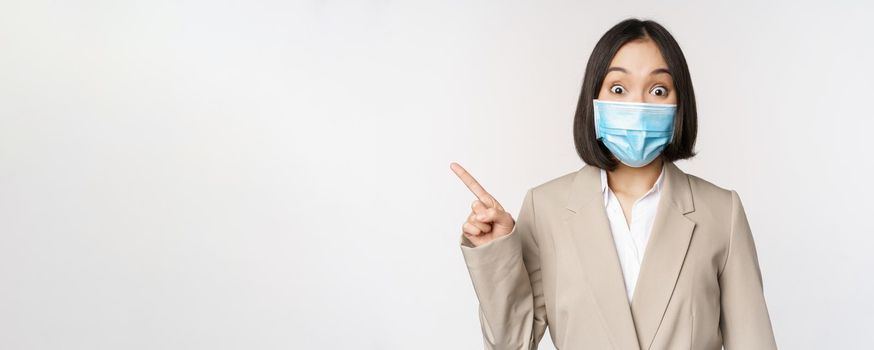 Coronavirus and work concept. Portrait of woman in medical face mask, pointing finger left, showing logo or banner, advertisement, white background.