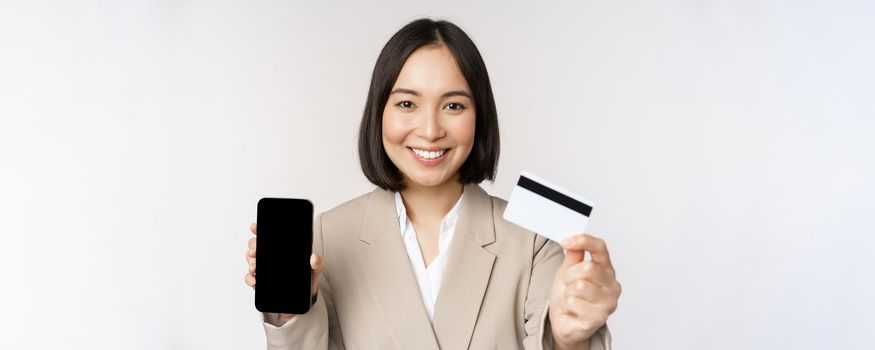 Smiling corporate woman in suit, showing mobile phone screen and app on mobile phone, smartphone screen, standing over white background.