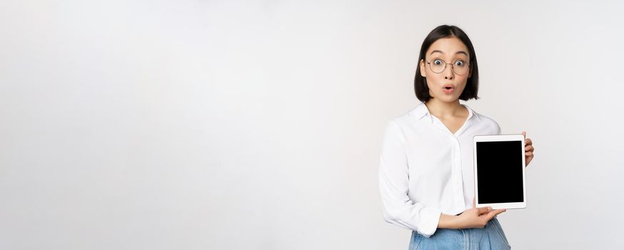 Enthusiastic asian woman, office worker in glasses showing digital tablet screen, demonstrating info on gadget display, standing over white background.