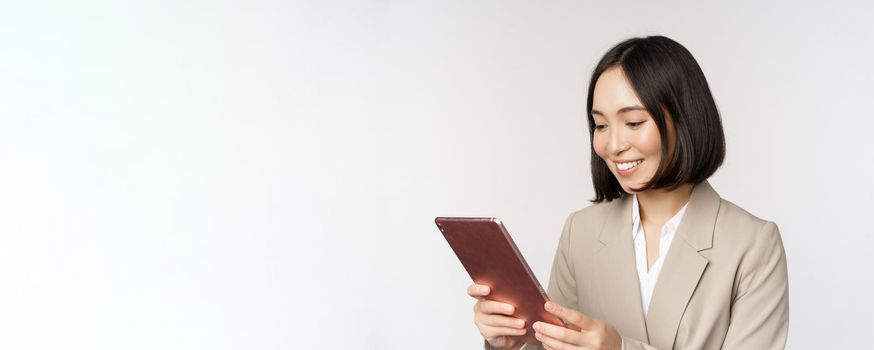 Image of asian businesswoman using digital tablet, looking at gadget and smiling, working, standing against white background.
