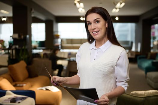 Attractive European woman, furniture store owner, successful sales rep and retail assistant holding a catalog with furniture and smiles looking at camera