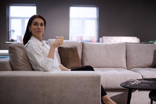 Relaxed beautiful European woman drinking coffee sitting on the comfortable sofa in the furniture store showroom. Interior design concept