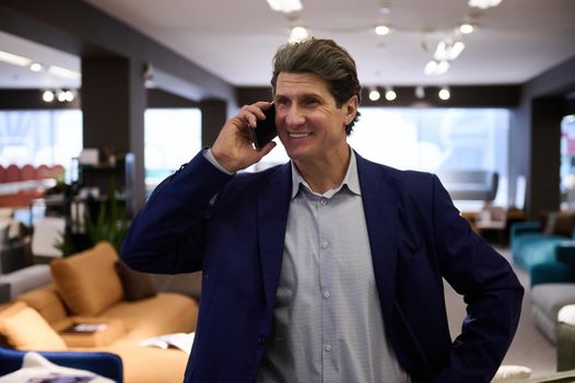 Middle aged Caucasian man talking on mobile phone while visiting a furniture store. Portrait of a confident businessman, entrepreneur, sales manager, retail assistant, interior designer