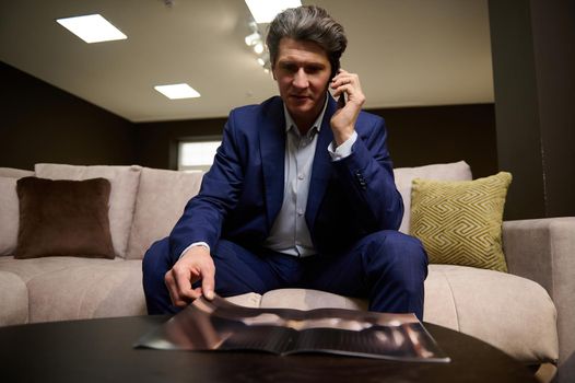 Mature man in business suit talking on the phone while sitting on a sofa and leafing through a catalog with assortment of upholstered furniture. Interior designer at work. Home improvement concept