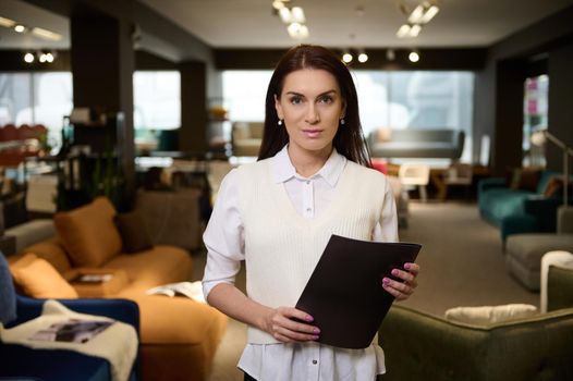 Confident female sales manager, successful retail assistant, beautiful brunette European woman standing with product catalog in furniture store exhibition center