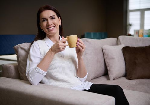 Beautiful serene European woman sitting on the sofa and feeling relaxed,enjoying a cup of coffee while picking a comfortable sofa in the furniture store showroom. Home interior design and improvement concept