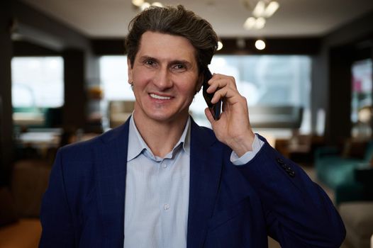 Close-up portrait of handsome mature Caucasian businessman, entrepreneur, interior designer in business suit smiles with beautiful toothy smile while talking on mobile phone and looking at camera