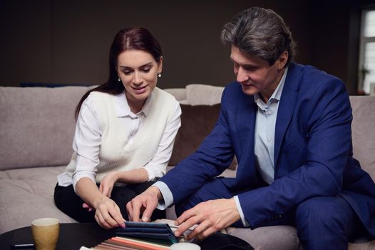 Relaxed middle aged couple picking color fabric samples for upholstery of the furniture, sitting on the sofa in the furniture store showroom