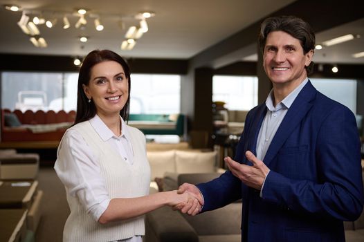 Mature sales representative retail manager and pleasant woman customer shake hands, smile looking at camera, standing together in a furniture store showroom. Furnishing and and interior design concept