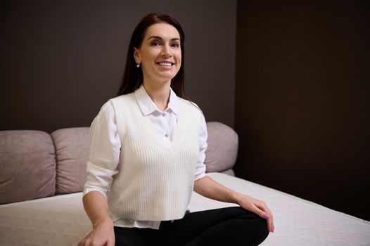 Attractive middle aged woman in casual wear sitting in lotus position on an orthopedic mattress and smiling looking at the camera while choosing a mattress for her bed in a furniture store showroom