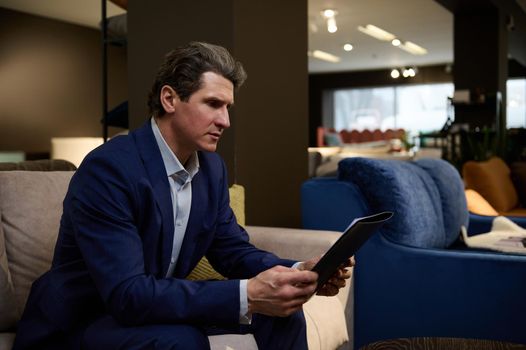 Handsome mature Caucasian man customer in business suit holding a catalog of upholstered furniture in the sofas exposition hall