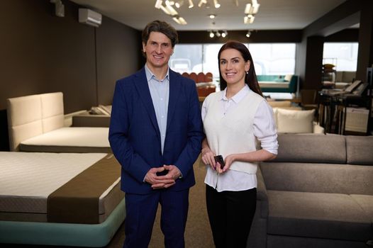 Successful diverse business partners- beautiful woman and handsome man, sales managers, assistants smile looking at camera, standing between beds with mattresses in a furniture store exhibition center