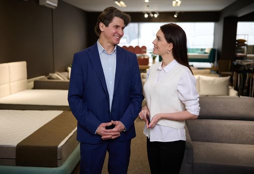 Beautiful European woman and handsome man- sales managers or happy middle aged married couple in casual clothes working or choosing bed and mattresses in the furniture store showroom