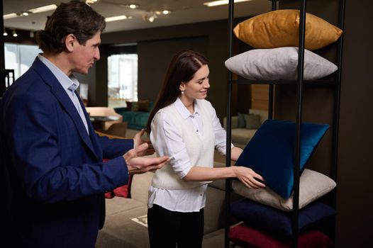 A charming retail assistant, sales manager and customer near a stand with stylish velour sofa pillows and cushions in a furniture store showroom. Exposition center with upholstered furniture