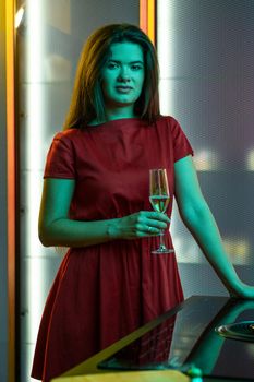 Young attractive brunette in red dress standing with glass of champagne in subdued color lighting in stylish home kitchen interior. House party concept