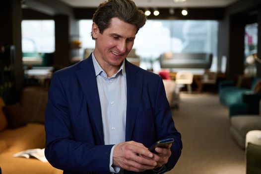 Handsome successful businessman, interior designer, furniture store owner typing text on his smartphone while working in the exhibition center of upholstered furniture