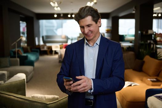 Handsome interior designer, businessman typing message on mobile phone, visiting a furniture store showroom. Home improvement and interior design concept.