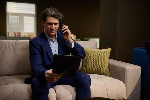 Handsome imposing mature man looking at catalog and talking on mobile phone while visiting a showroom. Retail assistant consulting clients and agreeing on the delivery time of upholstered furniture