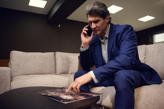 Handsome imposing mature Caucasian man, interior designer, successful businessman, entrepreneur, sales manager negotiating and advising clients on phone, looking furniture catalog in his cozy office