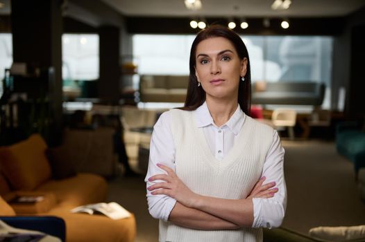 Confident middle aged business woman in casual clothes - successful sales assistant in the furniture store exhibition center, posing with crossed arms looking at camera