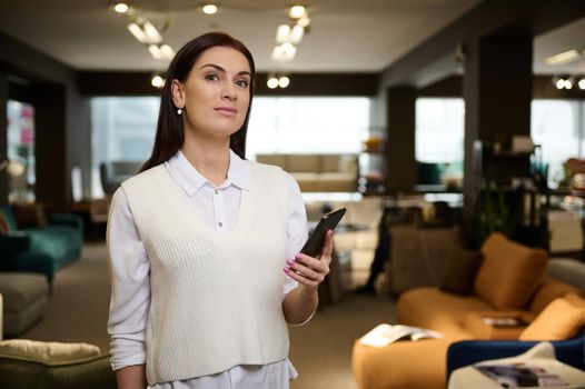 Attractive middle aged confident European woman in casual attire, sales manager, businesswoman, assistant standing with mobile phone in hand in the exhibition hall of upholstered furniture store