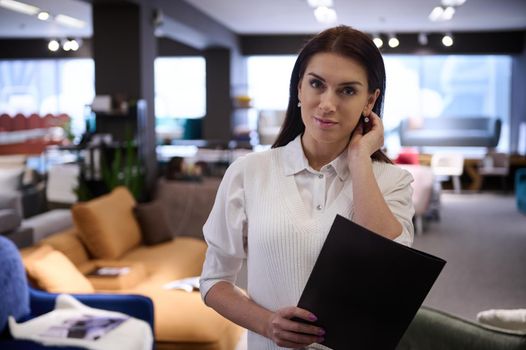 Female sales assistant, confident business woman with catalog in an exhibition hall of upholstered furniture store. Exposition of modern comfortable sofas, couches, soft beds and orthopedic mattresses