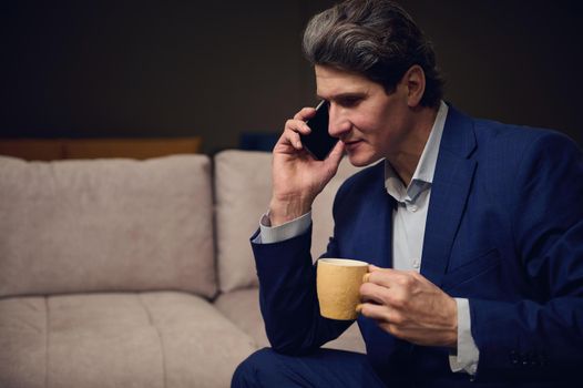 Handsome imposing mature man, interior designer, successful businessman, entrepreneur, sales manager negotiating and advising clients on the phone while having a coffee break in his cozy office