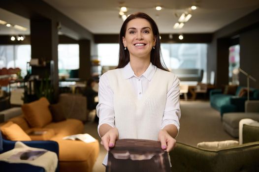 Pleasant pretty woman, retail assistant and sales rep showing catalog with upholstered furniture, standing in the exposition center and smiling with cheerful toothy smile looking at camera