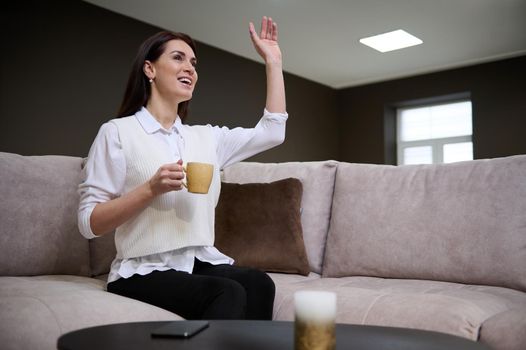 Attractive successful confident European business woman sitting with a cup of coffee on a sofa in the showroom of a furniture store and waving smiling greeting someone during a business meeting